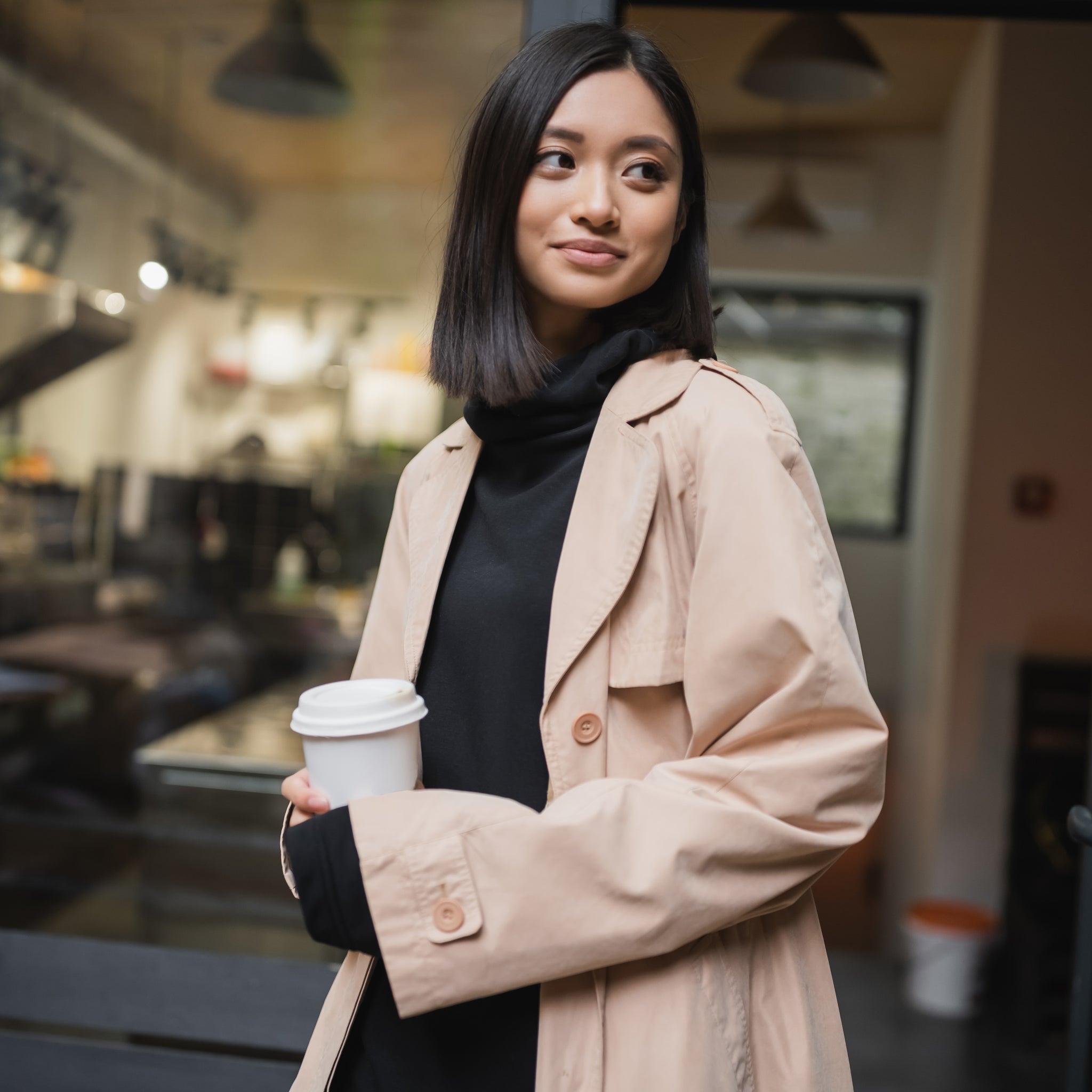 Woman in a beige coat holding a coffee cup outside a cafe.